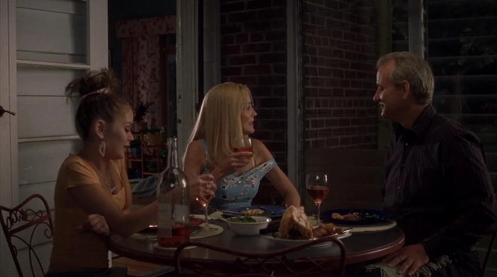 Broken Flowers movie still: medium — A dimly lit outdoor patio at night features a round table with dinner items. Three people sit at the table…