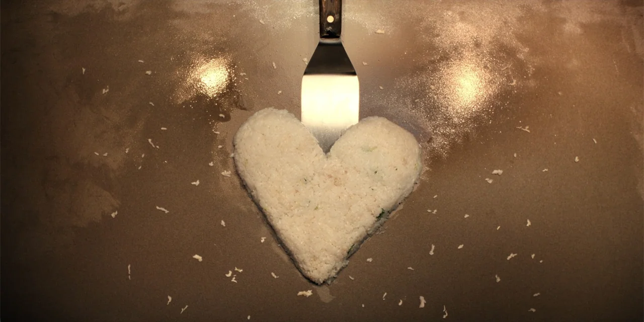 The Monkey (2025) movie still: closeup — A flat metallic cooking surface features a heart-shaped mound of white fried rice. A metal spatula rests…