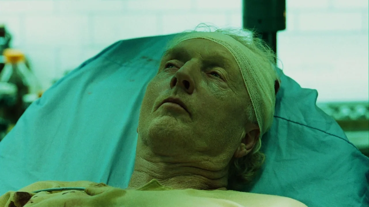 Saw III (2006) movie still: closeup — A stark hospital room features pale green walls and a blue medical bed cover. An elderly man with a head…