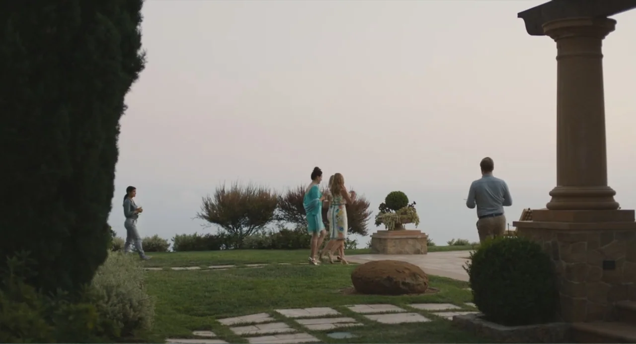 Beatriz at Dinner (2017) movie still: wide — An outdoor stone patio and green lawn overlook a distant, hazy landscape. Several people are walking…