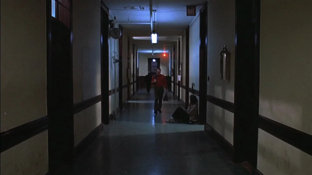 The Exorcist (1973) movie still: wide — A long, dimly lit building corridor features cream walls and dark wood trim. A man walks toward the camera…