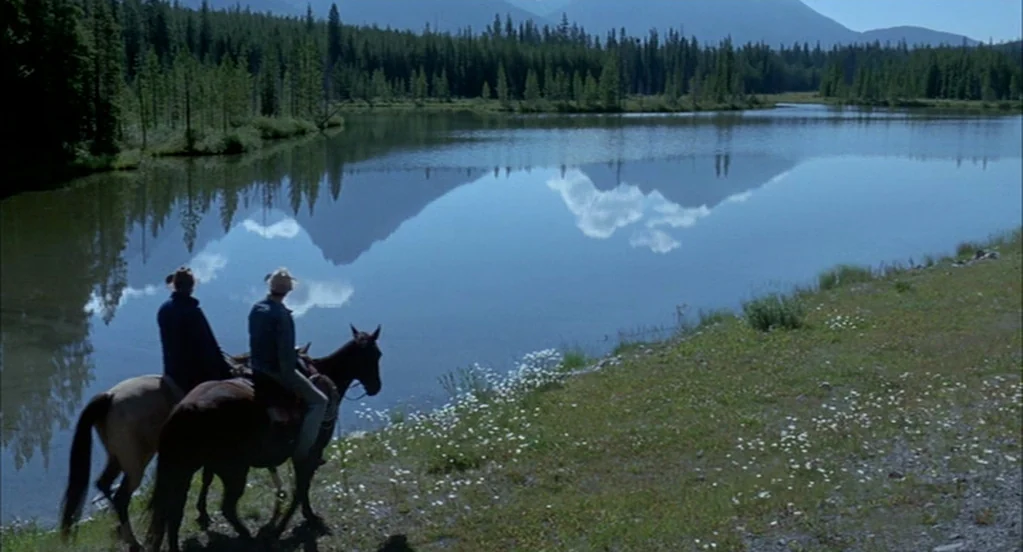 Brokeback Mountain (2005) movie still: wide — A calm, reflective lake is bordered by a pine tree forest and distant mountains. Two men sit on horses…