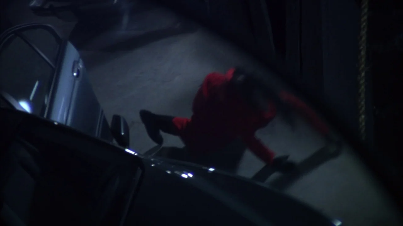 Saw movie still: wide — An asphalt ground surface at night with a car door open on the left. A person wearing a red jacket falls onto the ground in…