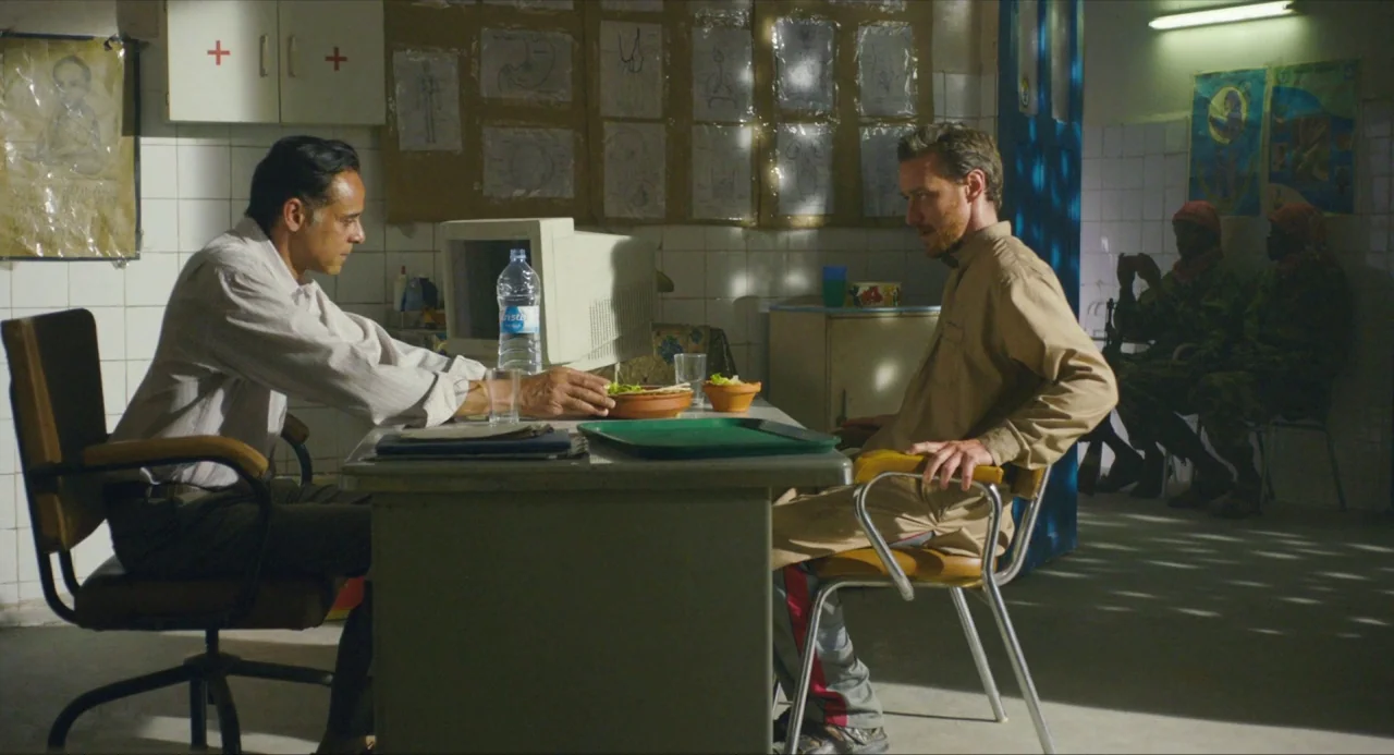 Submergence movie still: medium — A cluttered, sparsely furnished room features medical diagrams tacked onto the white tiled walls. Two men are…