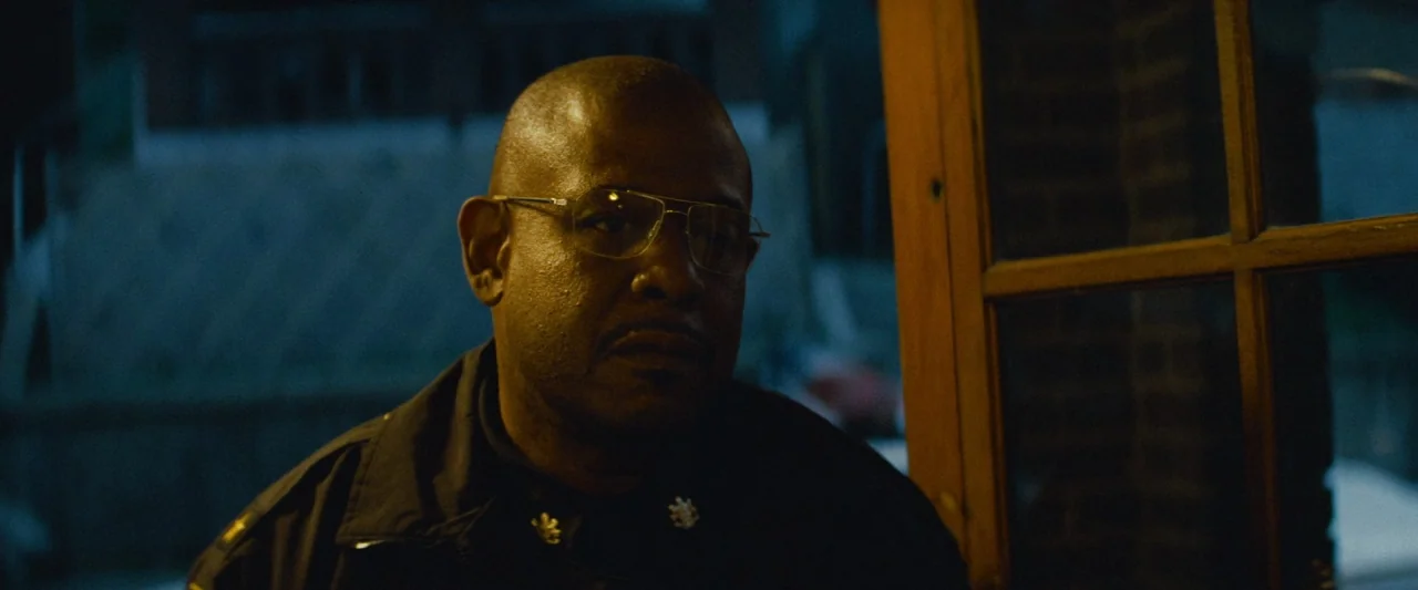 Out of the Furnace (2013) movie still: closeup — A man in a police uniform stands in the dark on an outdoor porch. A wooden door frame is visible to…