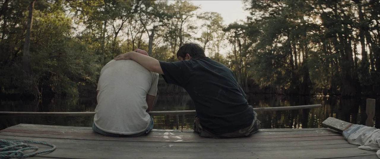 The Peanut Butter Falcon (2019) movie still: fullbody — A wooden pier sits on the surface of a dark river surrounded by trees. Two people sit side…