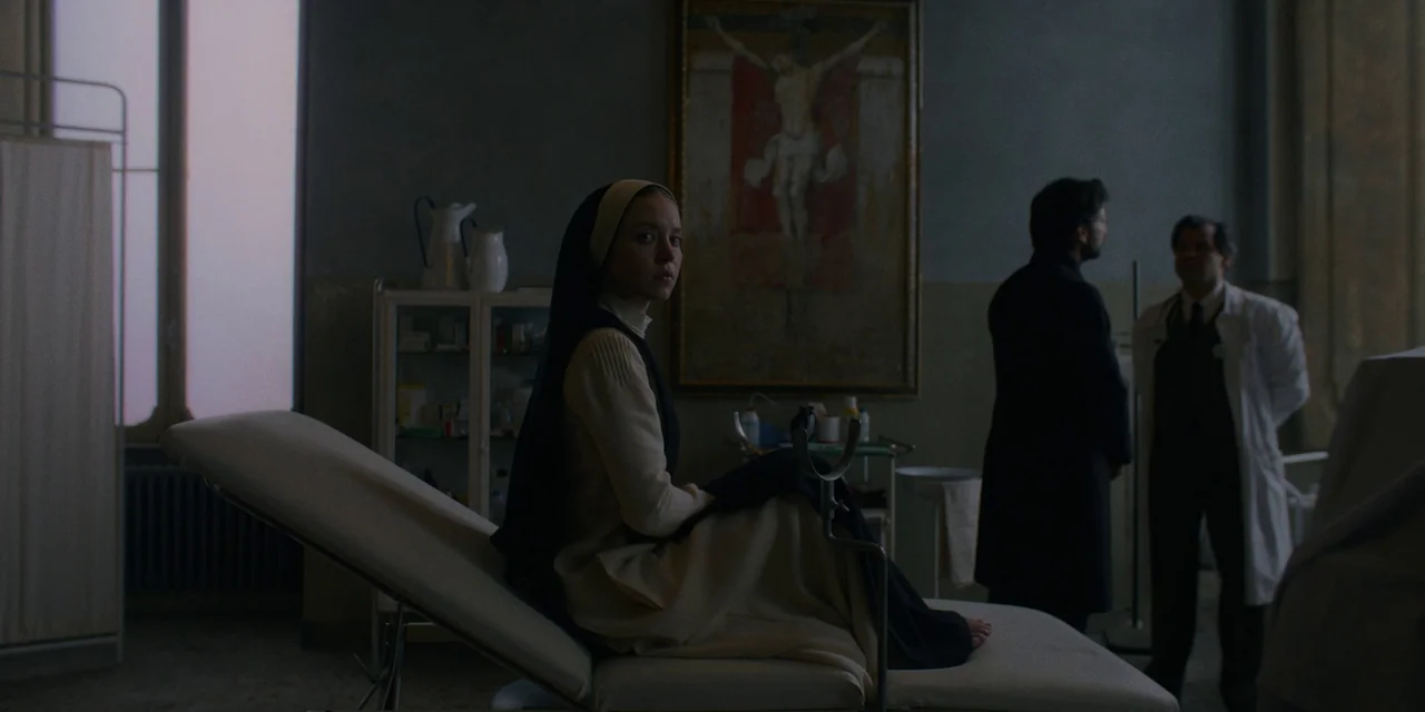 Immaculate (2024) movie still: medium — A dark examination room features a medical table and a framed painting of Jesus. A young woman in a…
