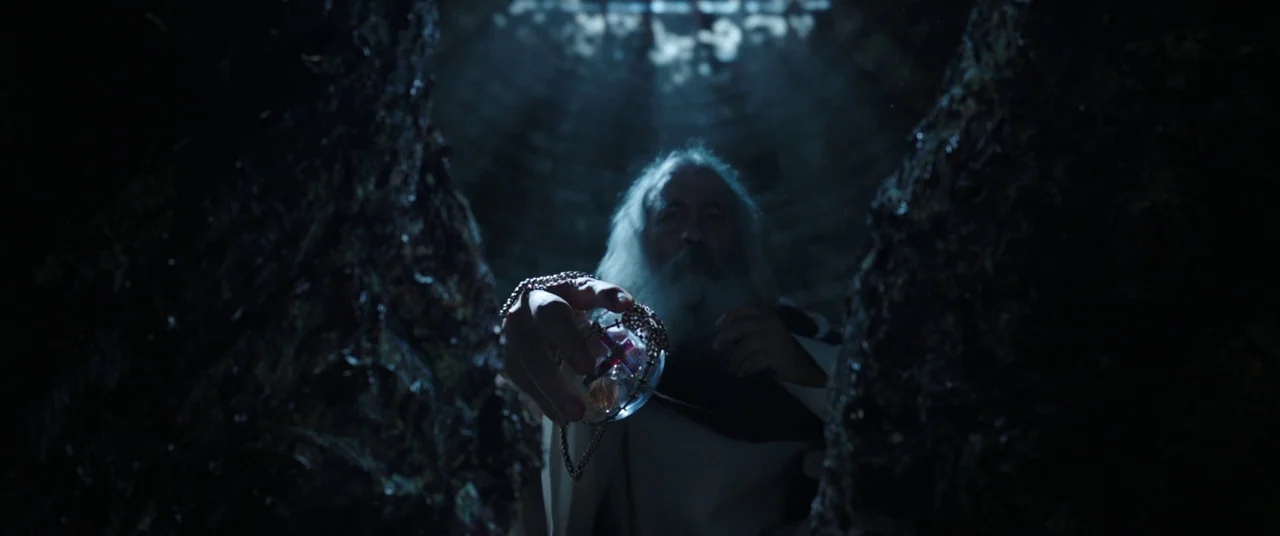The Nun (2018) movie still: medium — A dark stone interior features rough textures with a narrow shaft of light above. An elderly man in robes holds…