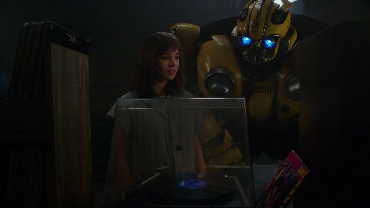 Bumblebee (2018) movie still: medium — A dimly lit industrial space contains a vintage record player and a tall robot. A teenage girl stands before…
