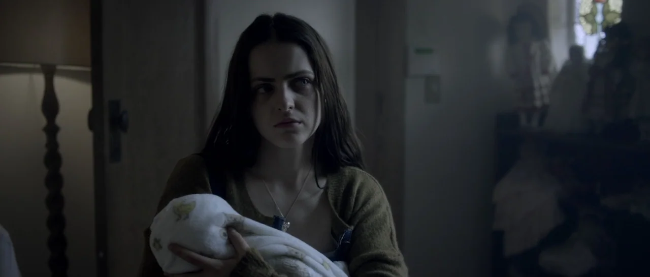 The Lullaby (2019) movie still: medium — A dimly lit room contains a young woman and an infant wrapped in white cloth. The woman looks away from the…