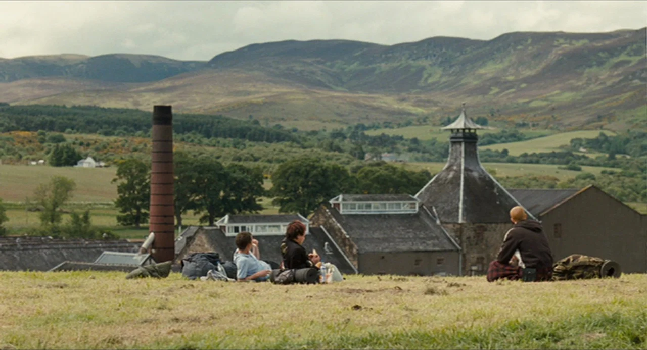 The Angel’s Share (2012) movie still: wide — A group of three people rests on a grassy ridge overlooking a rural site. Distant rolling mountains…