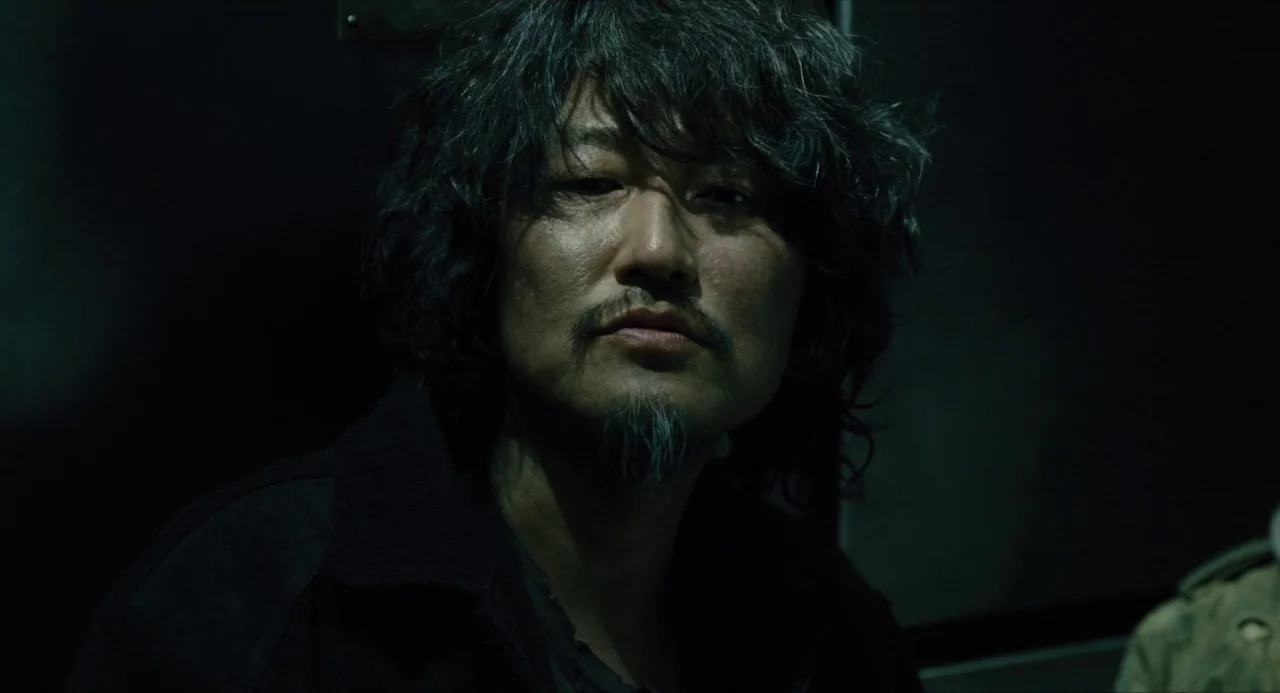Snowpiercer (2013) movie still: closeup — A man stands in a dark, confined space with minimal environmental detail visible. He looks straight ahead…