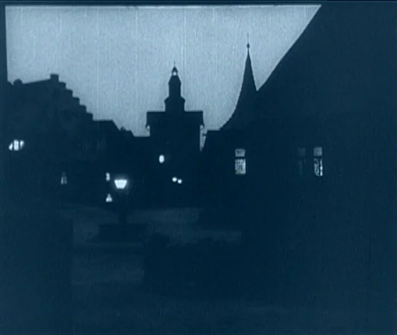 Destiny (1921) movie still: establishing — A dark, silhouetted view of a European town square with steep-roofed medieval architecture. A lone…