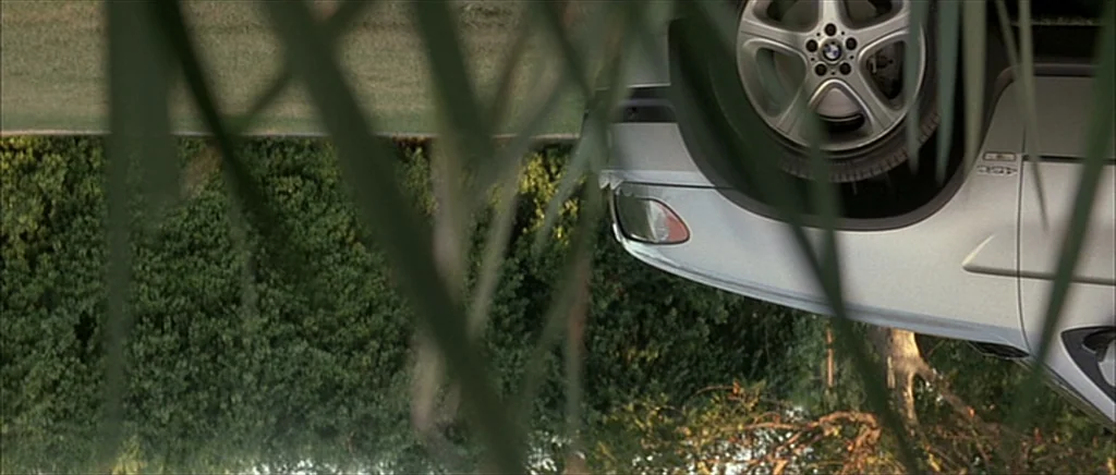 dutch angle shots example from I Heart Huckabees: A silver BMW SUV is viewed through foreground grass and shrubs on a roadside. The camera is rotated