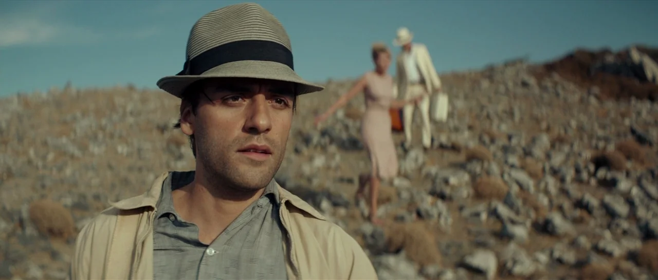 The Two Faces of January (2014) movie still: medium — A man in a hat stands in the foreground of a dry rocky hill. Two people walk in the background…
