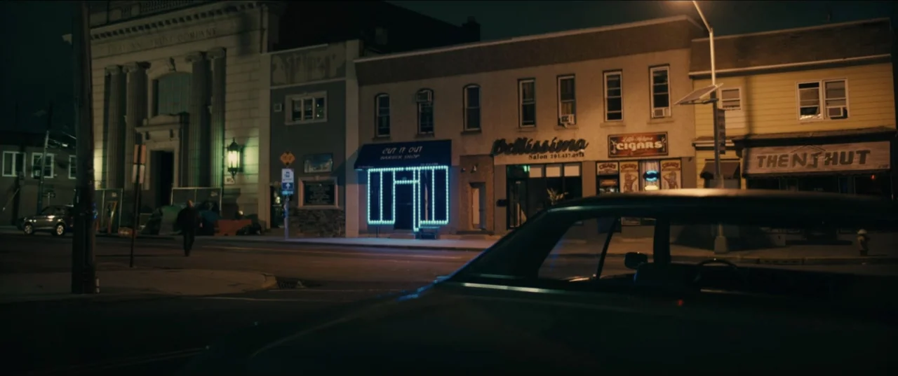 Guns Up (2025) movie still: wide — A nighttime city street features a row of small commercial storefronts and a bank. A parked vehicle occupies the…