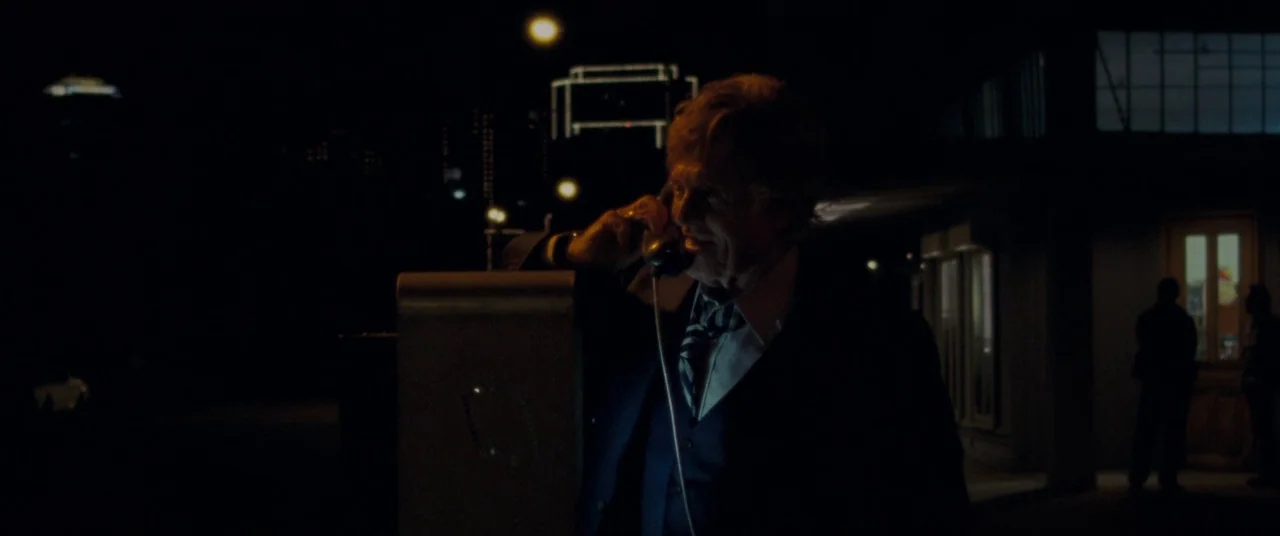 The Old Man & The Gun (2018) movie still: medium — A dark, nighttime exterior featuring a man standing alone at an outdoor payphone. The background…