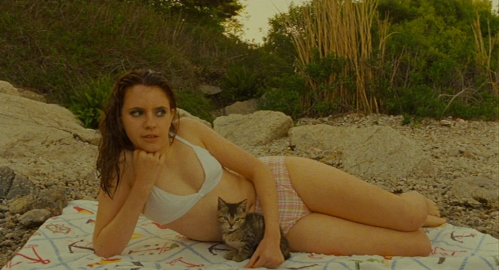 Moonrise Kingdom (2012) movie still: medium — A woman reclines on a patterned towel spread across a rocky shoreline. She rests on her arm beside a…