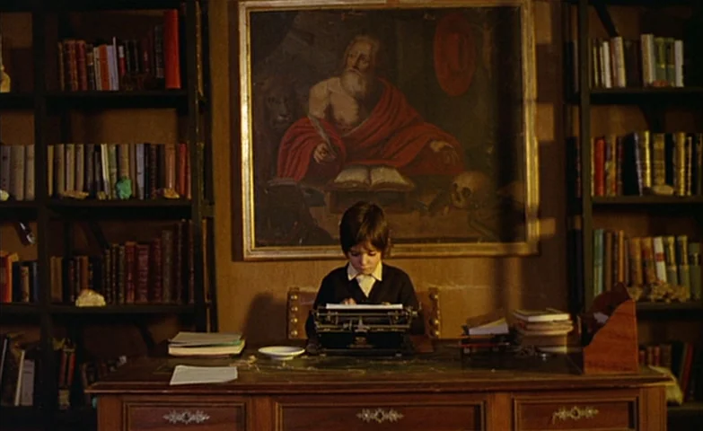 Spirit Of The Beehive (1973) movie still: medium — A dimly lit room contains walls lined with shelves of books and a painting. A young boy types on…