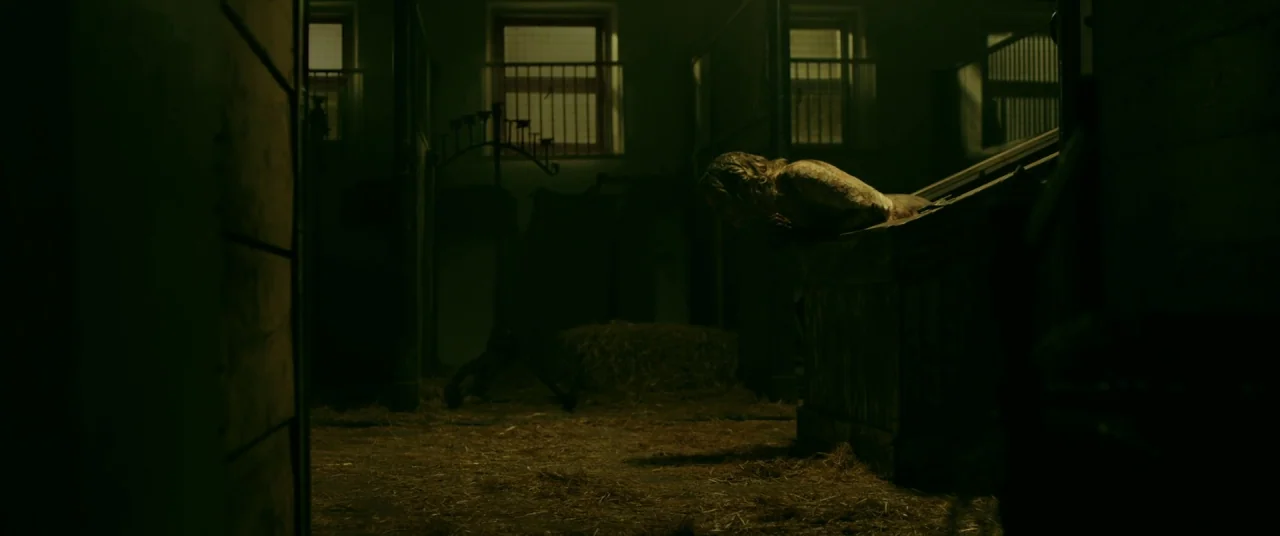 Ready or Not (2019) movie still: wide — A dark horse stable contains straw on the floor and wooden partitioned stalls. A young woman leans over the…