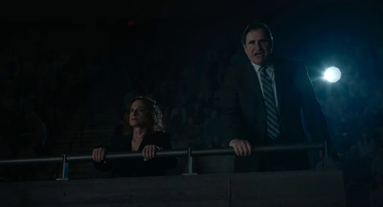 Beau Is Afraid (2023) movie still: medium — A dark, sparsely lit balcony overlooks a large, dimly lit sports arena. A man and a woman stand…