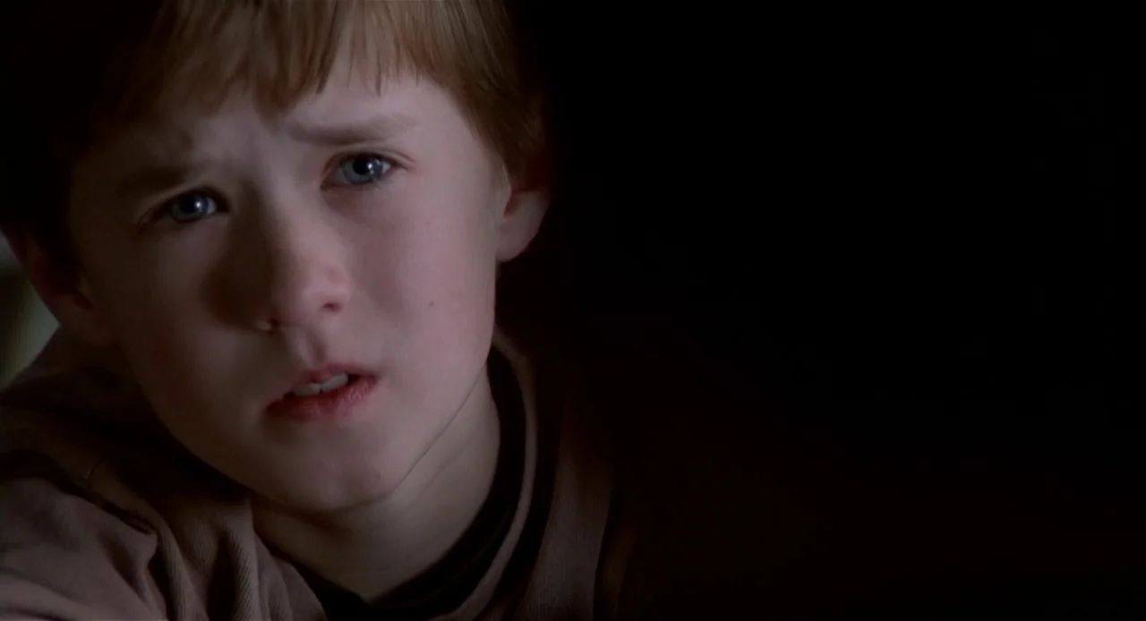 The Sixth Sense (1999) movie still: closeup — A dimly lit, dark space surrounds a young boy's face in close proximity. The child stares forward with…