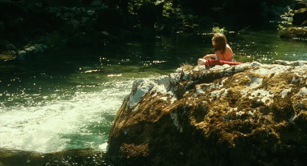 Things to Come (2016) movie still: wide — A clear, flowing creek with a large moss-covered rock in the foreground. A woman sits on the edge of the…