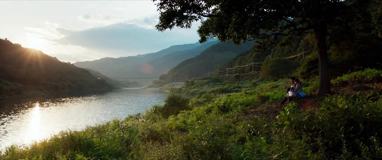 The Wailing (2016) movie still: wide — A lush riverbank features dense foliage and a large tree overlooking a wide river. Two people sit quietly on…