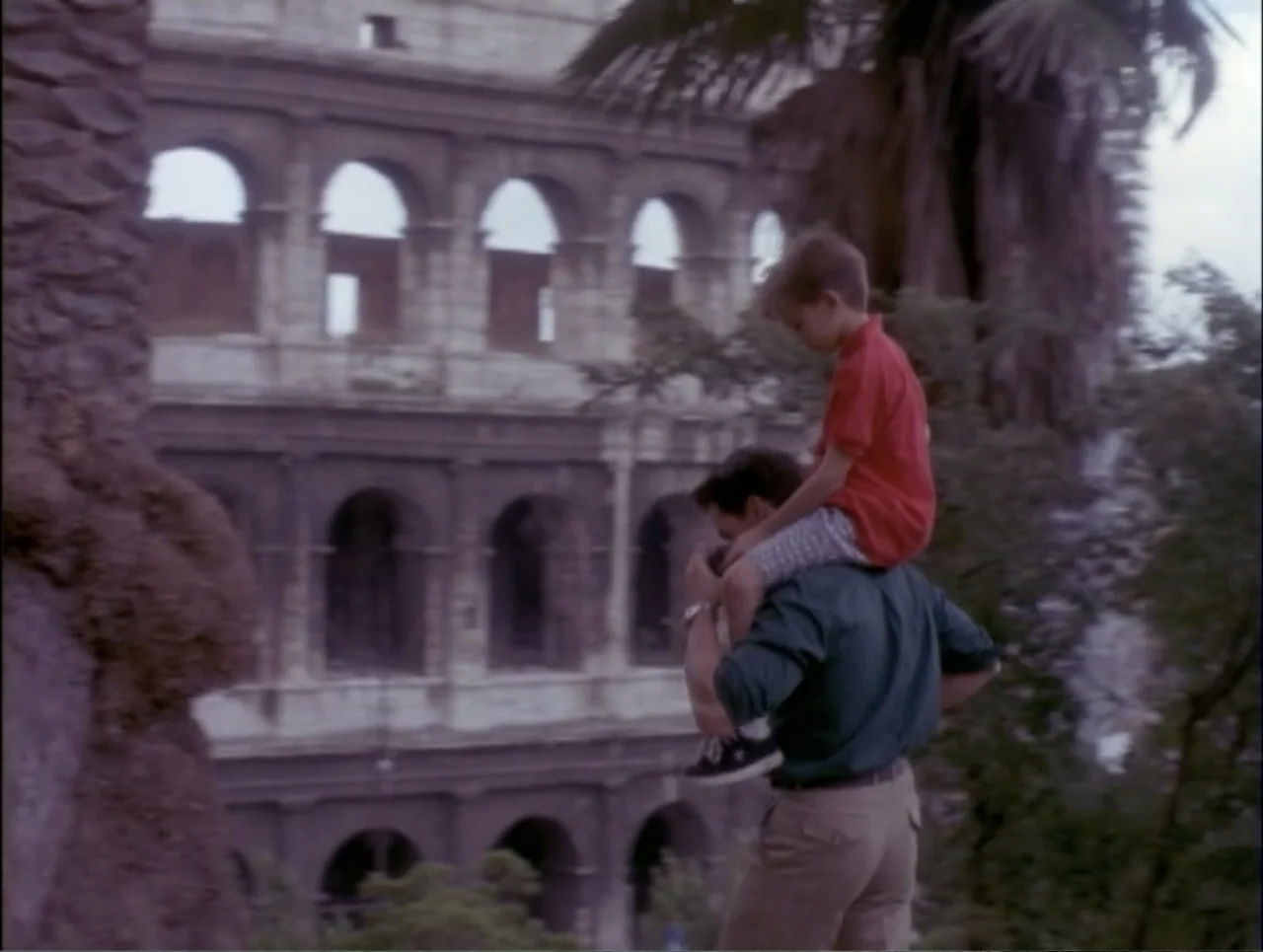 Code Name: Wolverine (1996) movie still: fullbody — A man carrying a boy on his shoulders stands before the large stone arches of the Colosseum. The…