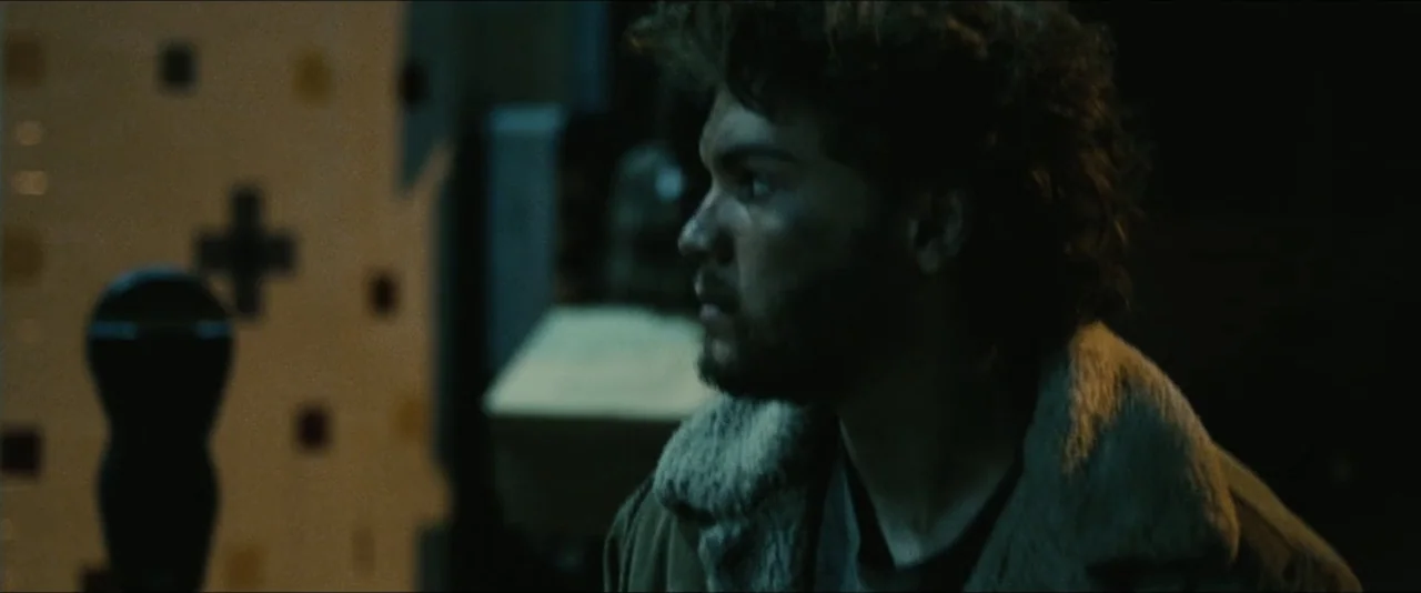 Into the Wild (2007) movie still: closeup — A dimly lit room with a patterned tiled wall in the background. A young man with a beard stands in the…