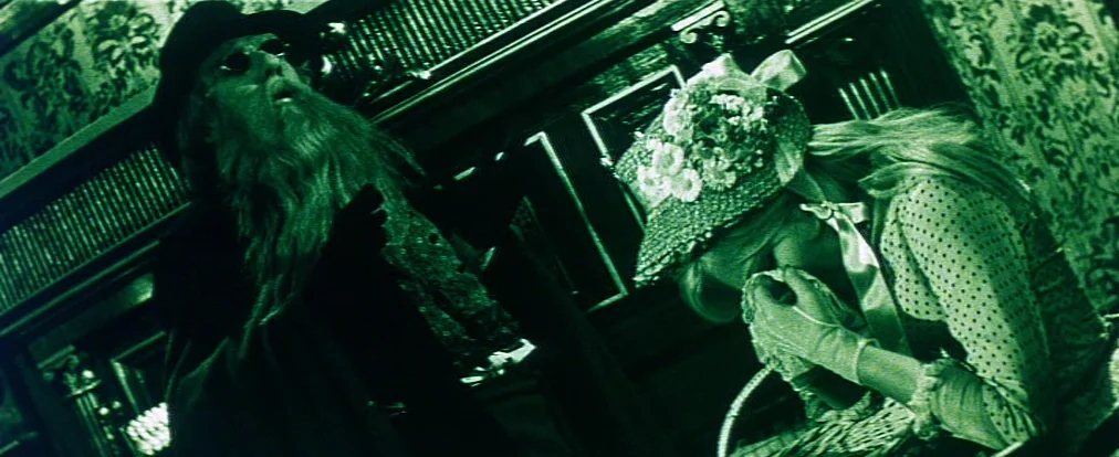 dutch angle shots example from Lemonade Joe: A dimly lit room features ornate Victorian-style wallpaper and heavy dark wood trim. An older man stands