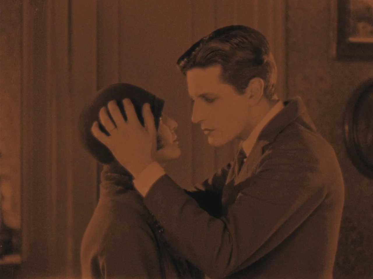 The Lodger (1927) movie still: medium — A dimly lit room with simple wall molding and a picture frame on the wall. A young man holds a woman's face…