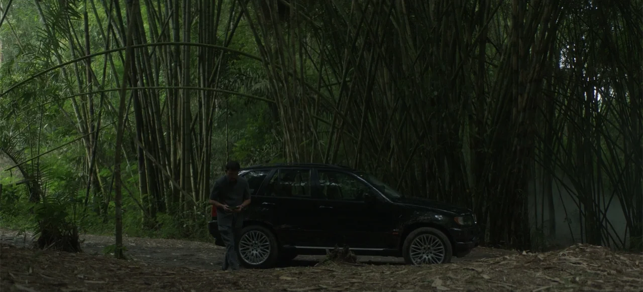 A Thousand Days (2023) movie still: wide — A dark SUV is parked on a dirt path surrounded by a tall bamboo grove. A man stands near the vehicle…