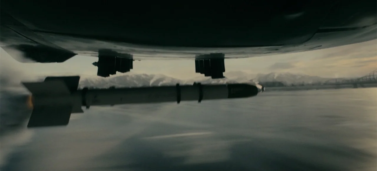 Devotion (2022) movie still: wide — A military missile is suspended under the wing of an aircraft flying over water. Snowy mountains are visible in…