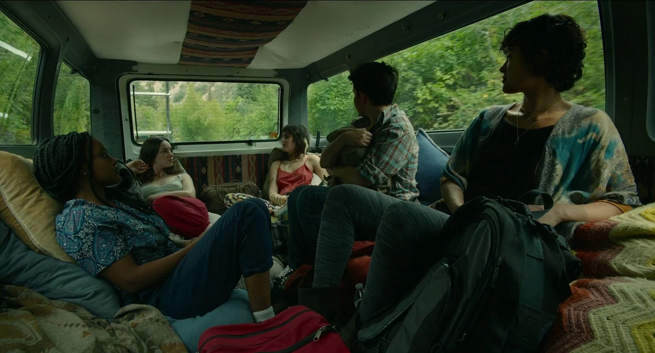Trim Season (2023) movie still: wide — The interior of a moving van is cluttered with pillows, blankets, and personal items. Five young adults sit…