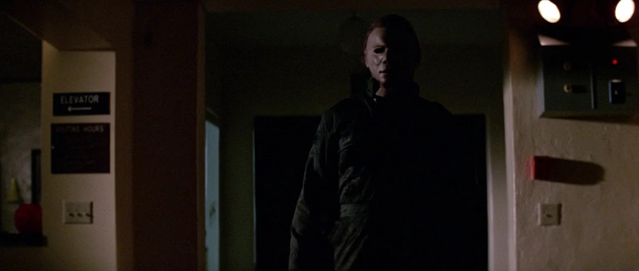 Halloween II (1981) movie still: medium — A dimly lit hospital corridor features dark walls and an elevator signage sign. A masked man stands…