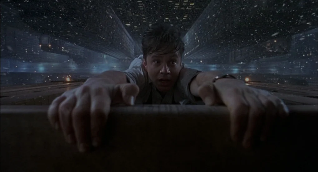 The Hudsucker Proxy (1994) movie still: closeup — A man hangs from the ledge of a rooftop at night overlooking a cityscape. The city appears to fold…