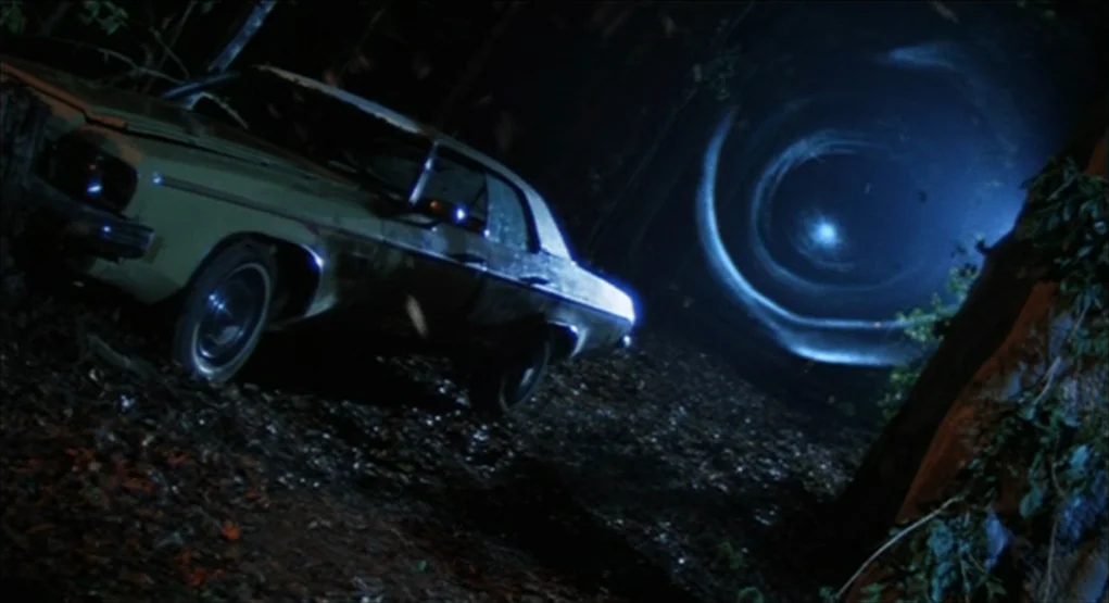 dutch angle shots example from Evil Dead II: A vintage sedan sits abandoned on a dark, tree-lined road at night. A bright circular blue light vortex s