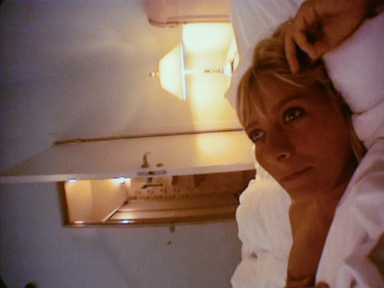 dutch angle shots example from Festen: A woman lies on white sheets inside a sparsely furnished room at night. A bright lamp glows in the background a