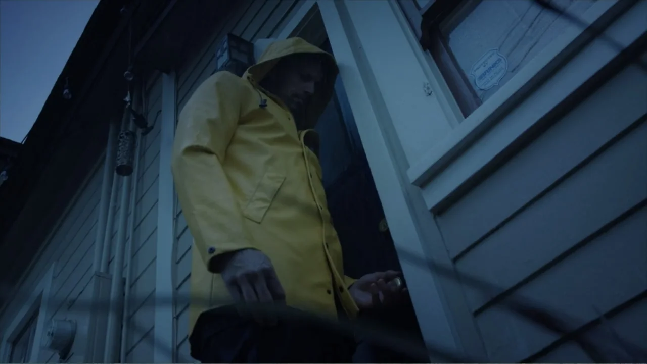 Storm Damage (2000) movie still: medium — A dark house exterior features a wooden porch at the onset of blue hour. A man in a bright yellow hooded…