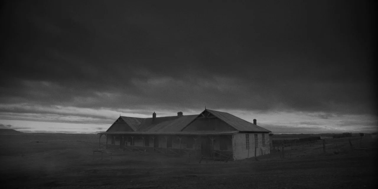 El Conde (2023) movie still: establishing — A low-slung, weathered wooden farmhouse stands alone in a vast, empty, barren landscape. The building is…