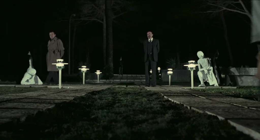 Todo Modo (1976) movie still: wide — A night scene in a park with paved paths flanked by small modern lights. Two men face each other from opposite…