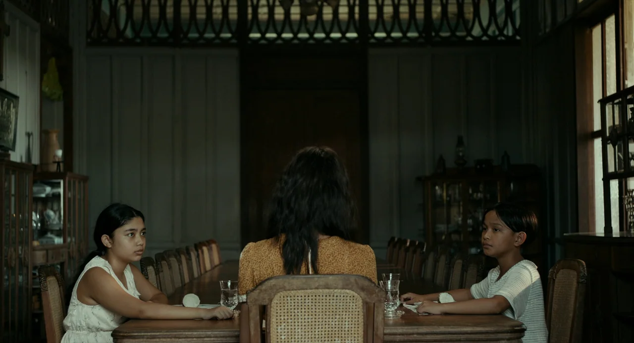 In My Mother’s Skin (2023) movie still: medium — A dark wooden dining area features a long table with cane-backed chairs and wood-paneled walls. An…