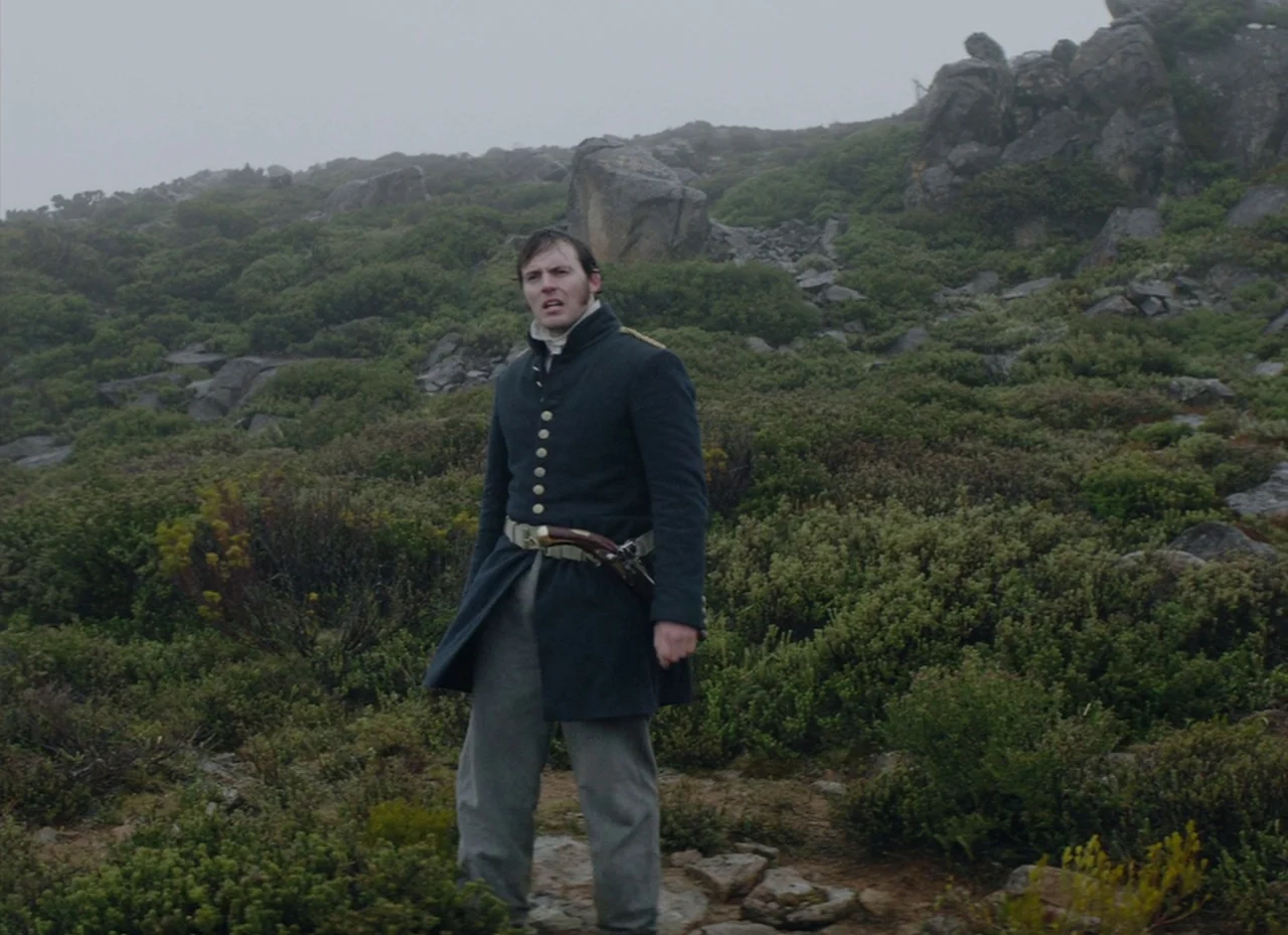 The Nightingale (2018) movie still: fullbody — A rocky, fog-covered mountain slope covered in low-lying green shrubs and vegetation. A man in…