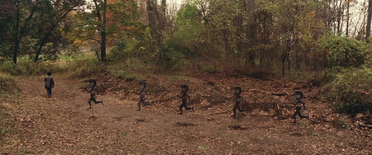 BlacKkKlansman (2018) movie still: wide — A leaf-strewn dirt path winds through a dense thicket of bare deciduous trees. A lone child walks away…