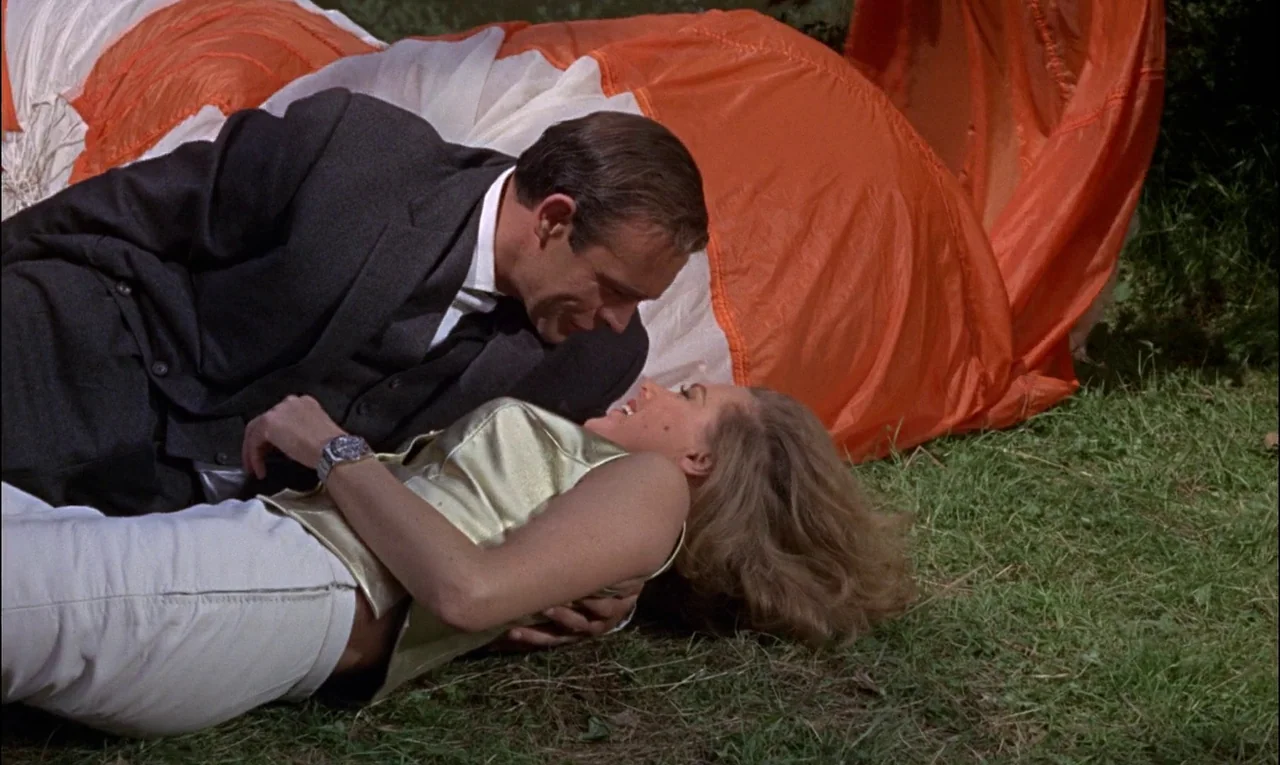 Goldfinger (1964) movie still: medium — A grassy field serves as the setting, featuring a large orange and white parachute. A man in a suit and a…
