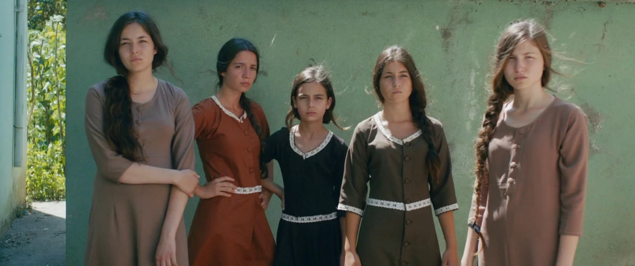 Mustang (2015) movie still: fullbody — Five young women stand in a row before a faded green concrete wall. They are wearing varied earth-toned…