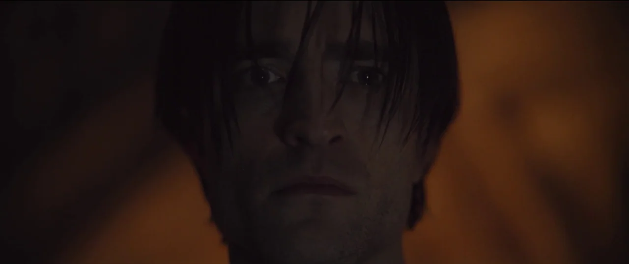 The Batman (2022) movie still: closeup — The man stands in a poorly lit space with indistinct dark surroundings. He gazes directly ahead with a…