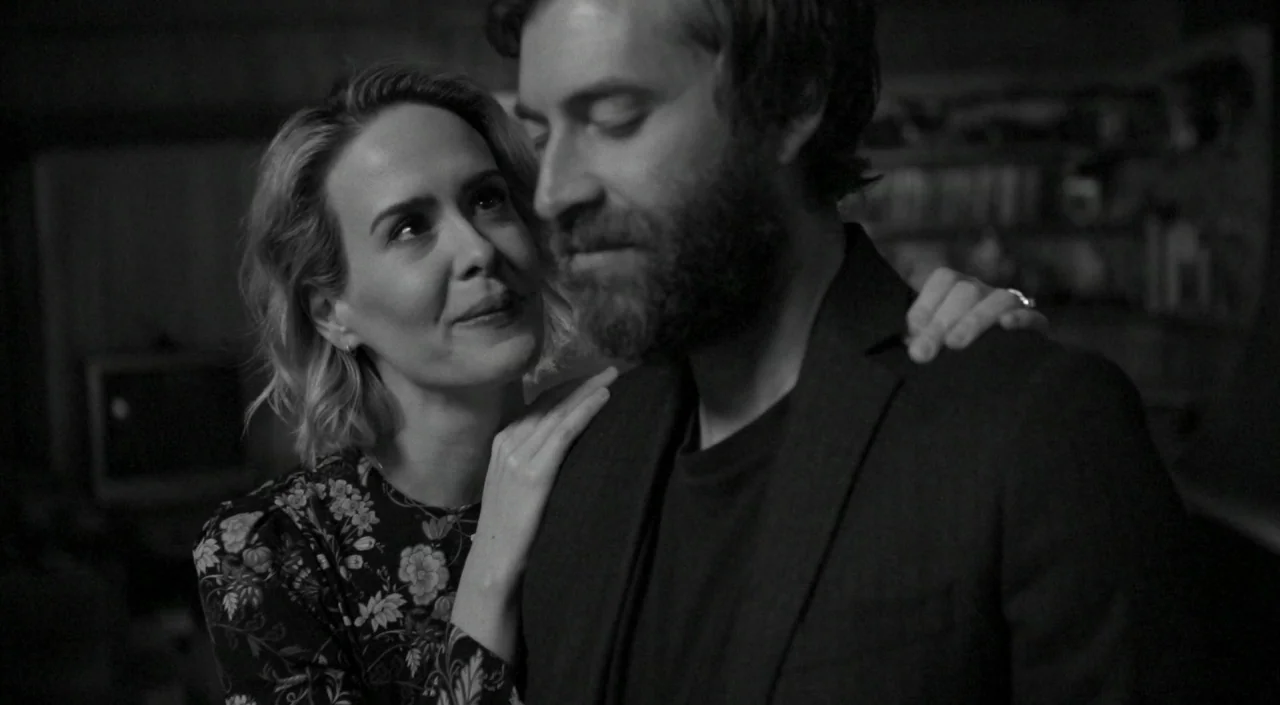 Blue Jay (2016) movie still: medium — A dark, dimly lit domestic space with blurred furniture in the background. A woman stands behind a man resting…