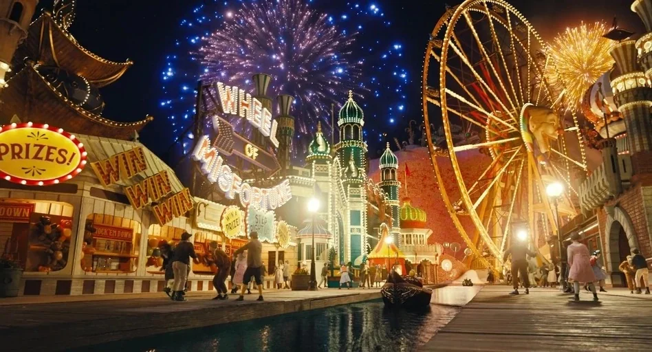 Pinocchio (2022) movie still: wide — An elaborate fantasy boardwalk with colorful stalls and a large Ferris wheel at night. Explosive fireworks…